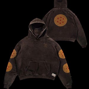 May include: Brown hoodie with a distressed finish, featuring a kangaroo pocket and a drawstring hood. The sleeves and back display circular patches with red stars. The initials "DB" are embroidered on the chest. A small tag is visible on the lower right.