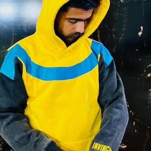 May include: Yellow hoodie with a blue stripe across the chest and gray sleeves. The hood is yellow, and the sleeves are gray with the word "INVINCIBLE" printed in yellow on the left sleeve. The hoodie is worn by a person.