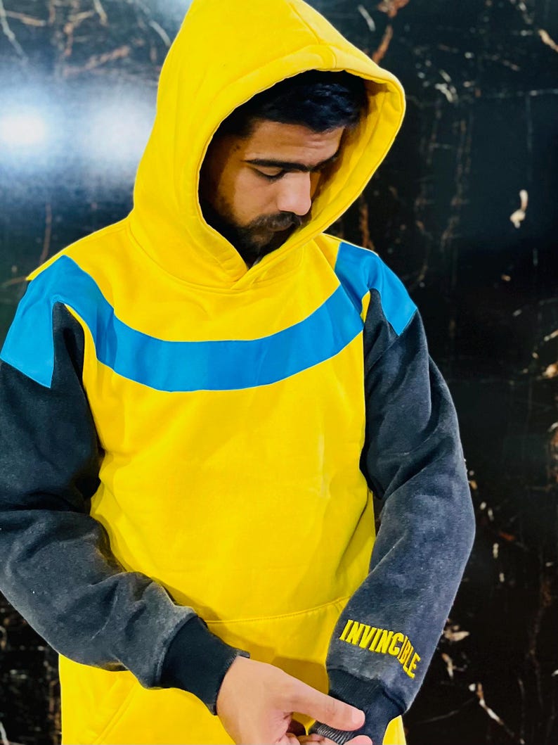 May include: Yellow hoodie with a blue stripe across the chest and gray sleeves. The hood is yellow, and the sleeves are gray with the word "INVINCIBLE" printed in yellow on the left sleeve. The hoodie is worn by a person.