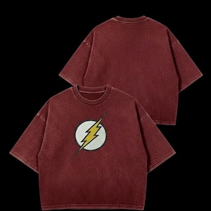 May include: Two maroon t-shirts with a faded appearance. The front shirt displays a white circle with a yellow lightning bolt design. The shirts have short sleeves and a crew neck. The background is black.