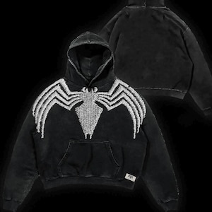 May include: Black hooded sweatshirt featuring a white embroidered spider design on the front. The hoodie has a kangaroo pocket and a drawstring hood. The design is a distressed style, with a faded appearance. The back of the hoodie is also visible.