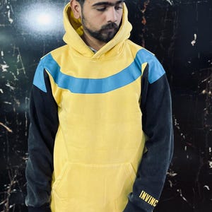 May include: Yellow hoodie with a blue stripe across the chest and black sleeves. The word "INVINC" is printed in gold on the left sleeve. The hoodie has a front pocket and a hood.