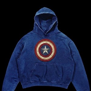 Puede incluir: Sudadera con capucha azul con un escudo del Capit&aacute;n Am&eacute;rica en el pecho. El escudo es rojo, blanco y azul con un borde dorado y una estrella plateada. La sudadera tiene un bolsillo de canguro.