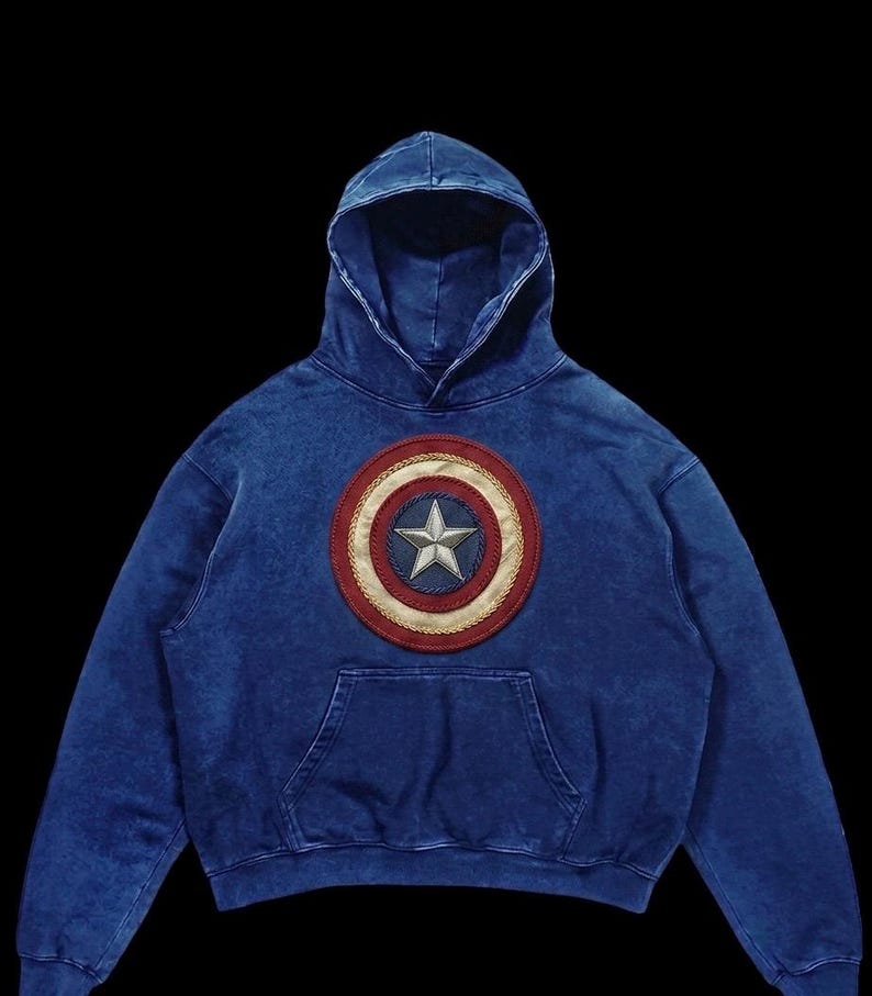 Puede incluir: Sudadera con capucha azul con un escudo del Capit&aacute;n Am&eacute;rica en el pecho. El escudo es rojo, blanco y azul con un borde dorado y una estrella plateada. La sudadera tiene un bolsillo de canguro.