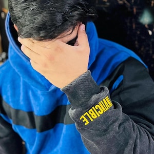 May include: Blue hoodie with black sleeves and a black stripe across the chest. The left sleeve cuff features the word "INVINCIBLE" in yellow lettering. The hoodie has a hood and a drawstring.