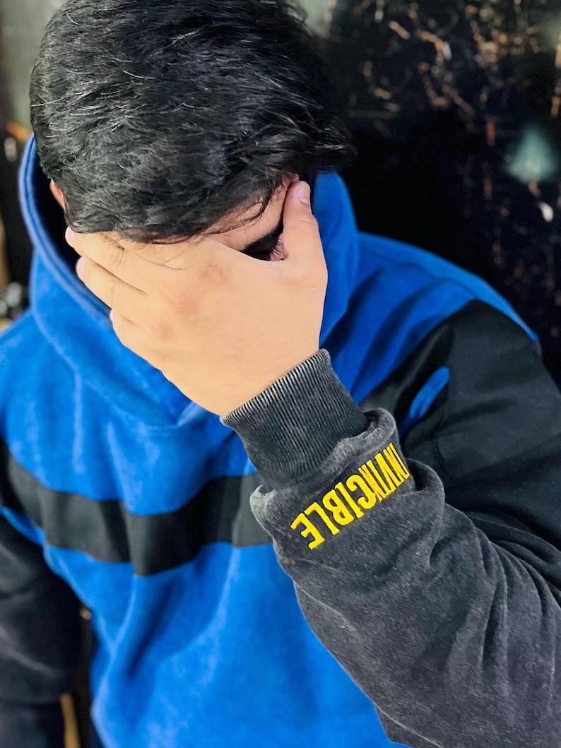 May include: Blue hoodie with black sleeves and a black stripe across the chest. The left sleeve cuff features the word "INVINCIBLE" in yellow lettering. The hoodie has a hood and a drawstring.