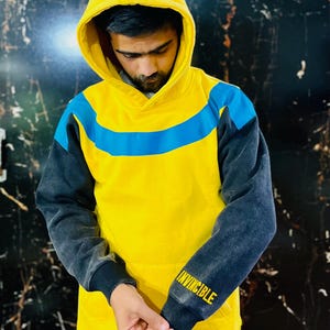 May include: Yellow hoodie with a blue stripe across the chest and dark gray sleeves. The word "INVINCIBLE" is printed in yellow on the left sleeve cuff. The hood is up, and the person is looking down.