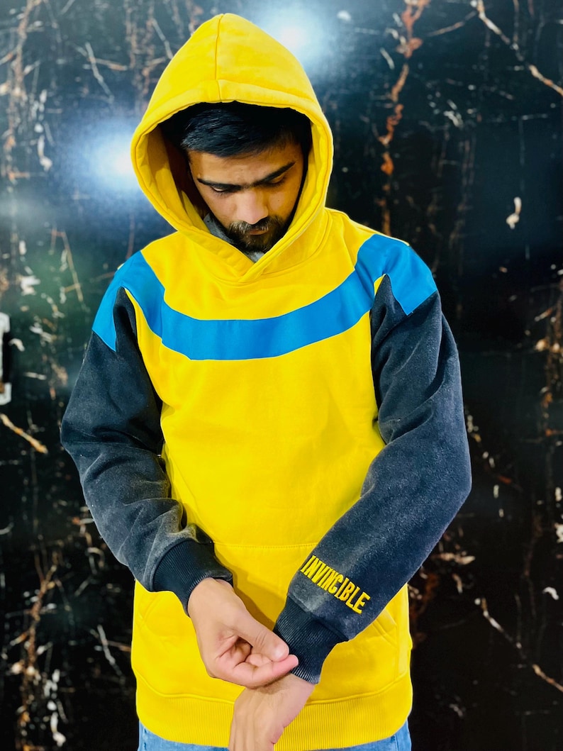 May include: Yellow hoodie with a blue stripe across the chest and dark gray sleeves. The word "INVINCIBLE" is printed in yellow on the left sleeve cuff. The hood is up, and the person is looking down.
