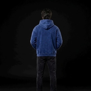 Puede incluir: Una persona con una sudadera con capucha azul y pantalones negros de pie sobre un fondo negro. La sudadera tiene una apariencia texturizada. La persona est&aacute; de espaldas a la c&aacute;mara.