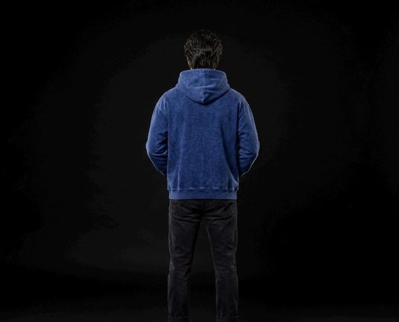 Puede incluir: Una persona con una sudadera con capucha azul y pantalones negros de pie sobre un fondo negro. La sudadera tiene una apariencia texturizada. La persona est&aacute; de espaldas a la c&aacute;mara.