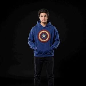 Puede incluir: Una sudadera con capucha azul con el emblema del escudo del Capit&aacute;n Am&eacute;rica. El escudo es un dise&ntilde;o circular con c&iacute;rculos conc&eacute;ntricos rojos, blancos y azules y una estrella blanca en el centro. La persona lleva pantalones negros.