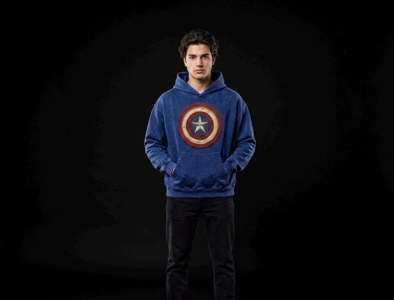 Puede incluir: Una sudadera con capucha azul con el emblema del escudo del Capit&aacute;n Am&eacute;rica. El escudo es un dise&ntilde;o circular con c&iacute;rculos conc&eacute;ntricos rojos, blancos y azules y una estrella blanca en el centro. La persona lleva pantalones negros.
