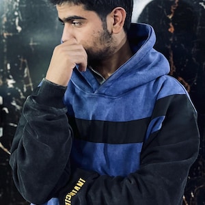 May include: A blue and black hoodie with a black stripe across the chest. The sleeves are black, and the left sleeve has the word "INVINCIBLE" printed in gold. The hoodie has a hood and is worn by a person.