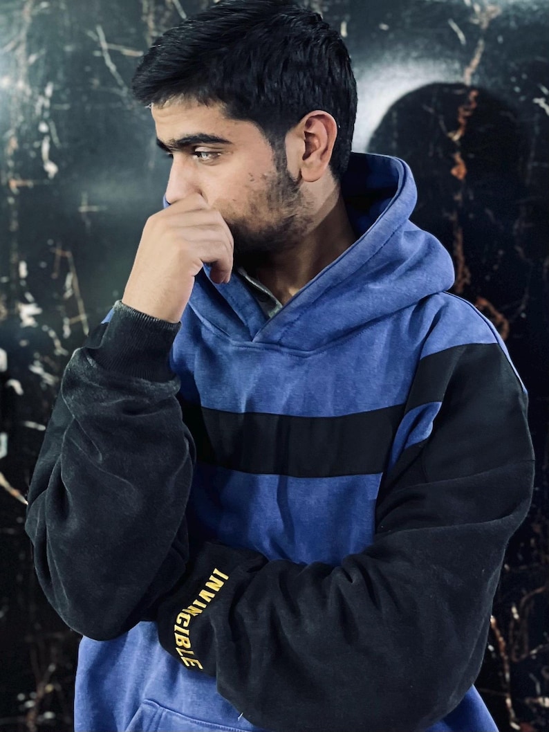 May include: A blue and black hoodie with a black stripe across the chest. The sleeves are black, and the left sleeve has the word "INVINCIBLE" printed in gold. The hoodie has a hood and is worn by a person.