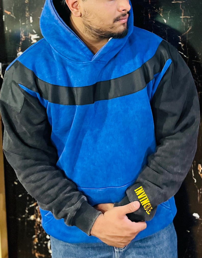 May include: Blue hoodie with black sleeves and a black stripe across the chest. The left sleeve cuff has the word "INVINCIBLE" in yellow lettering. The hoodie has a drawstring hood and a kangaroo pocket.