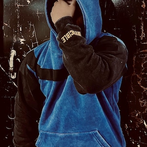 May include: A blue and black hoodie with the word "INVINCIBLE" on the sleeve. The hoodie has a front pocket and a drawstring hood. The blue fabric has a textured, faded appearance.