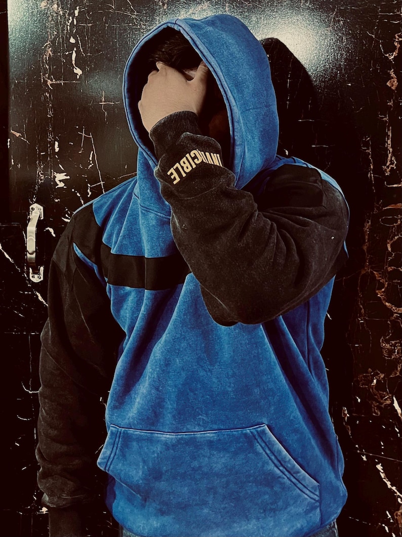 May include: A blue and black hoodie with the word "INVINCIBLE" on the sleeve. The hoodie has a front pocket and a drawstring hood. The blue fabric has a textured, faded appearance.