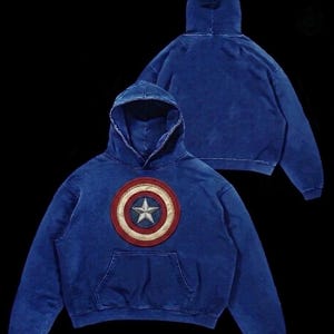 Puede incluir: Sudadera con capucha azul con un escudo del Capit&aacute;n Am&eacute;rica. El escudo es un dise&ntilde;o circular con una estrella blanca en el centro, rodeada de c&iacute;rculos conc&eacute;ntricos rojos, blancos y azules. La sudadera tiene un bolsillo delantero y una capucha con cord&oacute;n.