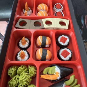 May include: A red bento box filled with various sushi pieces. The sushi includes nigiri, maki rolls, and temaki, with ingredients like salmon, shrimp, and avocado. The box also contains wasabi and pickled ginger.