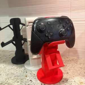 May include: A black Nintendo Switch Pro controller sits on a red 3D-printed stand. The stand has a circular base and an adjustable arm. Two other stands, one black and one white, are in the background.