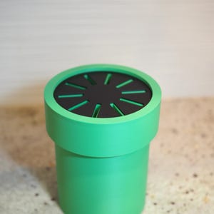 Op de afbeelding: Een groene cilindrische container met een zwarte bovenkant met radiale uitsparingen. De container heeft een gladde, matte afwerking en een afgeronde rand. Het ontwerp doet denken aan een videogamepijp.