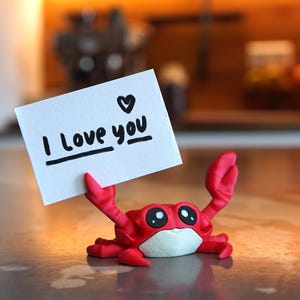 May include: A red crab figurine holding a white card with the handwritten message "I Love you" and a heart symbol. The crab has large, expressive eyes and is posed on a surface. The background is blurred, suggesting a shallow depth of field.