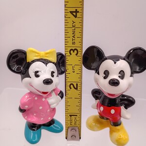 K&ouml;nnte beinhalten: Zwei Keramikfiguren von Mickey und Minnie Mouse. Minnie tr&auml;gt ein rosa Kleid mit wei&szlig;en Punkten, eine gelbe Schleife und blaue Schuhe. Mickey tr&auml;gt rote Shorts, wei&szlig;e Handschuhe und gelbe Schuhe. Die Figuren sind etwa 7,6 cm hoch.