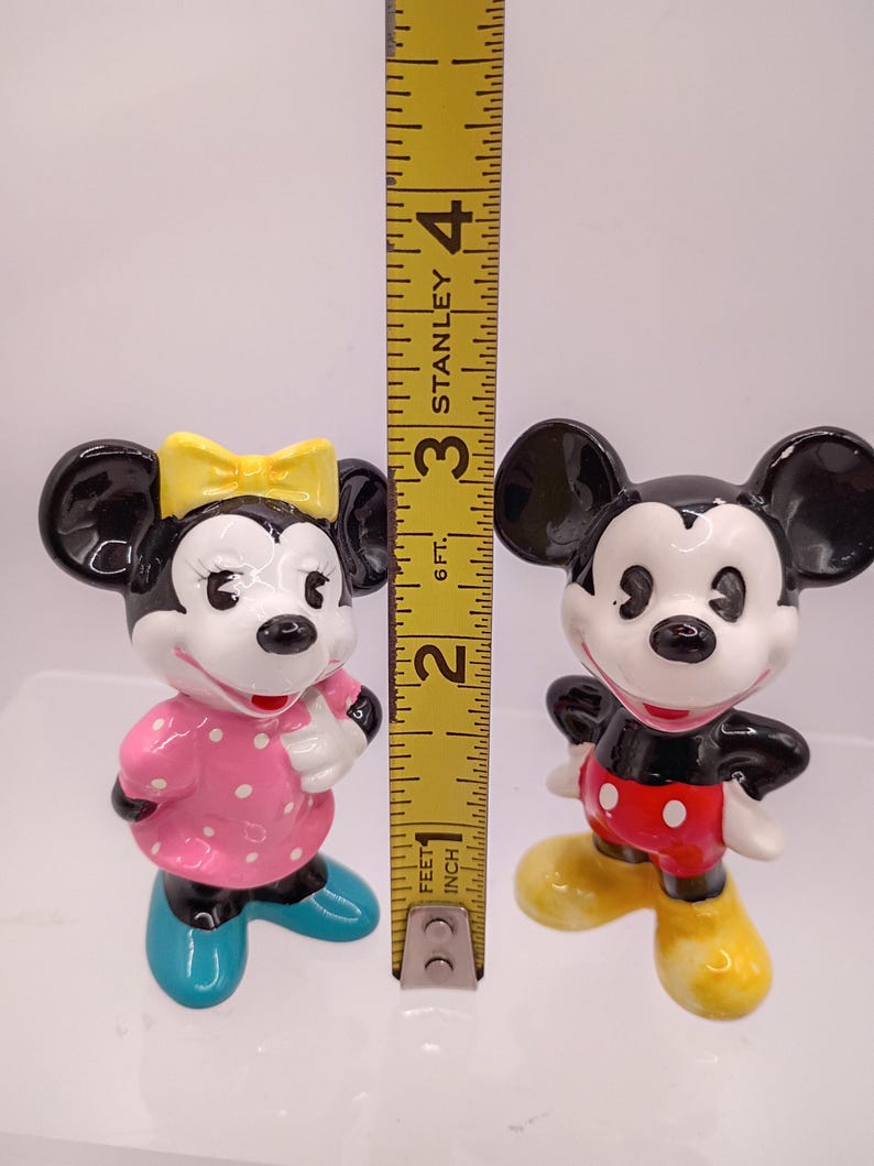 K&ouml;nnte beinhalten: Zwei Keramikfiguren von Mickey und Minnie Mouse. Minnie tr&auml;gt ein rosa Kleid mit wei&szlig;en Punkten, eine gelbe Schleife und blaue Schuhe. Mickey tr&auml;gt rote Shorts, wei&szlig;e Handschuhe und gelbe Schuhe. Die Figuren sind etwa 7,6 cm hoch.