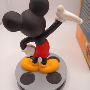 Puede incluir: Una figura de Mickey Mouse en una pose cl&aacute;sica, con orejas y cabeza negras, pantalones cortos rojos, zapatos amarillos y guantes blancos. La figura est&aacute; sobre una base de carrete de pel&iacute;cula gris. Una caja con texto es visible.