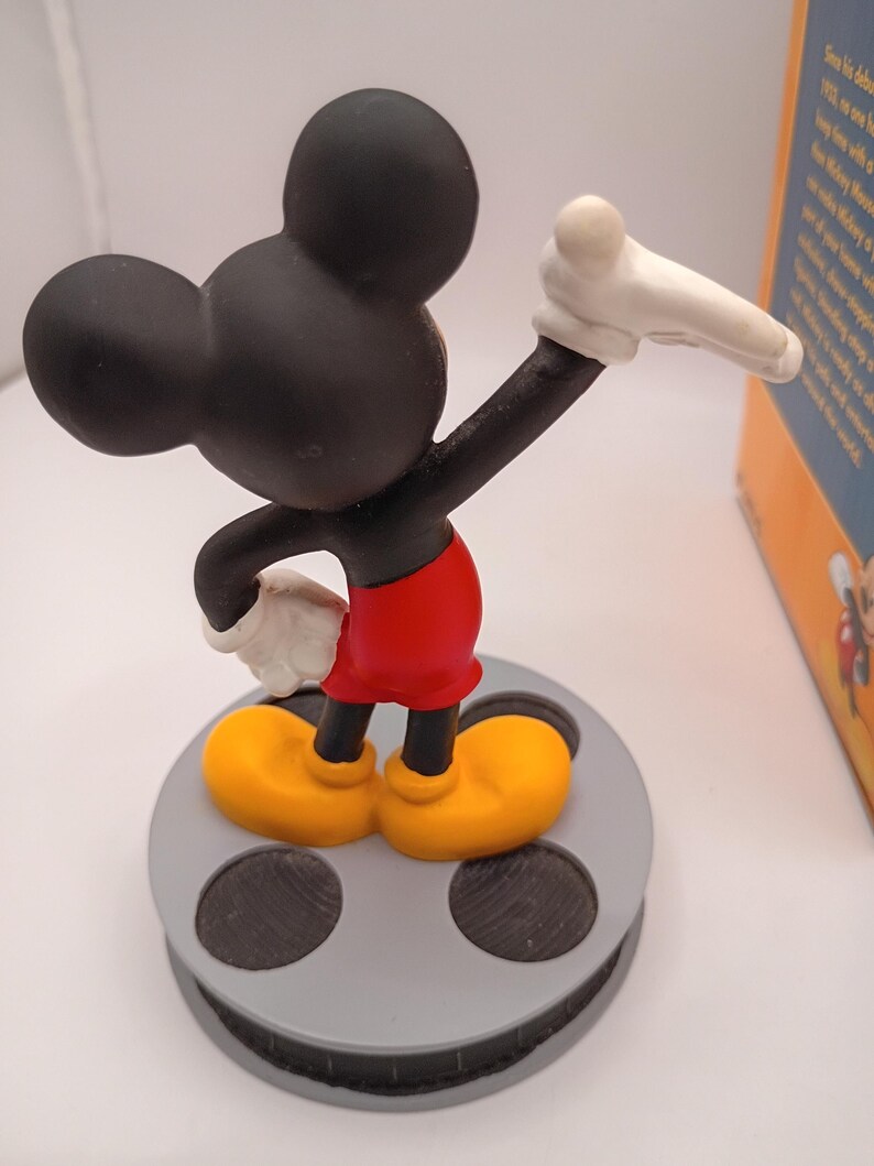 Puede incluir: Una figura de Mickey Mouse en una pose cl&aacute;sica, con orejas y cabeza negras, pantalones cortos rojos, zapatos amarillos y guantes blancos. La figura est&aacute; sobre una base de carrete de pel&iacute;cula gris. Una caja con texto es visible.