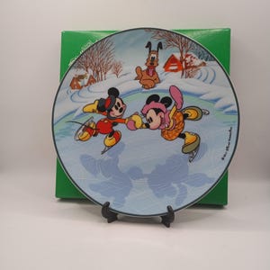 May include: A decorative plate featuring Mickey and Minnie Mouse ice skating on a frozen pond, with Pluto in the background. The plate has a winter scene with snow-covered ground, trees, and small houses. The plate is displayed on a black stand.