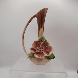 May include: A ceramic vase with a curved handle and a large, three-dimensional flower. The vase is primarily beige with a brown top and handle. The flower is a mix of pink and brown with green leaves.