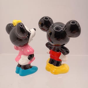 K&ouml;nnte beinhalten: Zwei Keramikfiguren von Micky und Minnie Maus. Micky tr&auml;gt ein rotes Hemd, gelbe Schuhe und schwarze Shorts. Minnie hat ein rosa Kleid mit wei&szlig;en Punkten, blaue Schuhe und eine gelbe Schleife. Beide haben schwarze Ohren.