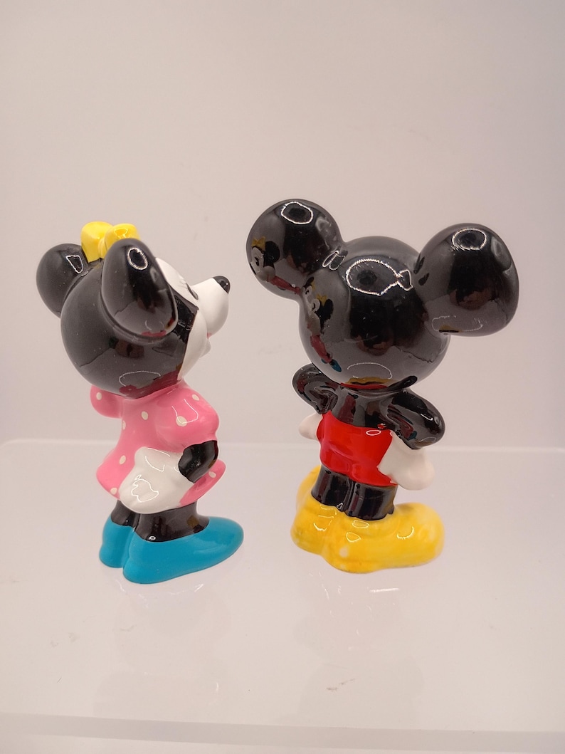 K&ouml;nnte beinhalten: Zwei Keramikfiguren von Micky und Minnie Maus. Micky tr&auml;gt ein rotes Hemd, gelbe Schuhe und schwarze Shorts. Minnie hat ein rosa Kleid mit wei&szlig;en Punkten, blaue Schuhe und eine gelbe Schleife. Beide haben schwarze Ohren.