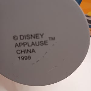 Puede incluir: Primer plano de un objeto gris y circular con el texto "&copy; DISNEY APPLAUSE TM CHINA 1999" impreso. El objeto es probablemente un art&iacute;culo de colecci&oacute;n o parte de un producto m&aacute;s grande.