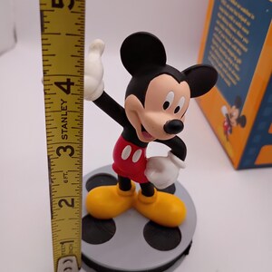 Puede incluir: Una figura de Mickey Mouse, de pie sobre una base de carrete de pel&iacute;cula gris. Mickey lleva pantalones cortos rojos, zapatos amarillos y guantes blancos. La figura mide aproximadamente 10 cm de altura, medida con una cinta m&eacute;trica amarilla Stanley.