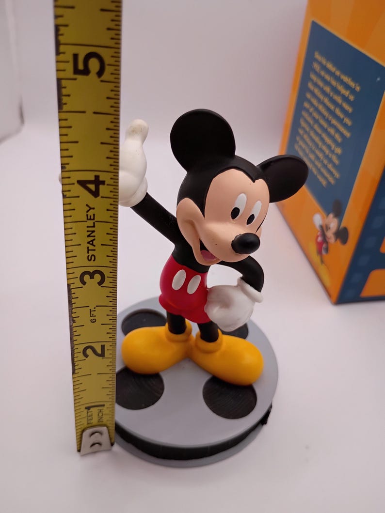 Puede incluir: Una figura de Mickey Mouse, de pie sobre una base de carrete de pel&iacute;cula gris. Mickey lleva pantalones cortos rojos, zapatos amarillos y guantes blancos. La figura mide aproximadamente 10 cm de altura, medida con una cinta m&eacute;trica amarilla Stanley.
