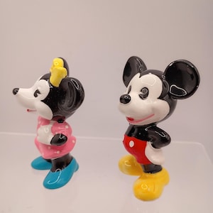 K&ouml;nnte beinhalten: Zwei Keramikfiguren von Mickey und Minnie Mouse. Mickey tr&auml;gt rote Shorts, gelbe Schuhe und wei&szlig;e Handschuhe. Minnie tr&auml;gt ein rosa Kleid mit wei&szlig;en Punkten, blaue Schuhe und eine gelbe Schleife.