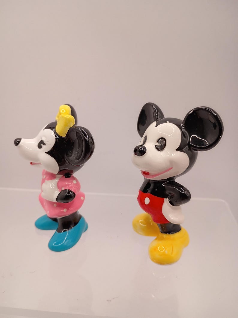 K&ouml;nnte beinhalten: Zwei Keramikfiguren von Mickey und Minnie Mouse. Mickey tr&auml;gt rote Shorts, gelbe Schuhe und wei&szlig;e Handschuhe. Minnie tr&auml;gt ein rosa Kleid mit wei&szlig;en Punkten, blaue Schuhe und eine gelbe Schleife.