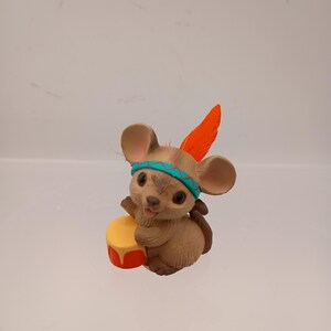 May include: A brown mouse figurine with a turquoise headband and an orange feather. The mouse is holding a yellow drum with red accents. The figurine has a detailed, textured appearance and is set against a plain background.