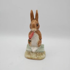 May include: A brown ceramic figurine of a rabbit sitting on a green bench with a carrot in its mouth. The rabbit is wearing a white shirt and has brown fur. The bench is on a base of brown grass.
