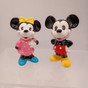 K&ouml;nnte beinhalten: Zwei Keramikfiguren von Mickey und Minnie Mouse. Mickey tr&auml;gt rote Shorts und gelbe Schuhe. Minnie tr&auml;gt ein rosa Kleid mit wei&szlig;en Punkten, eine gelbe Schleife und blaue Schuhe. Beide haben schwarze Ohren und wei&szlig;e Gesichter.