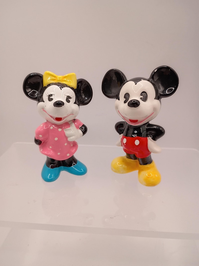 K&ouml;nnte beinhalten: Zwei Keramikfiguren von Mickey und Minnie Mouse. Mickey tr&auml;gt rote Shorts und gelbe Schuhe. Minnie tr&auml;gt ein rosa Kleid mit wei&szlig;en Punkten, eine gelbe Schleife und blaue Schuhe. Beide haben schwarze Ohren und wei&szlig;e Gesichter.