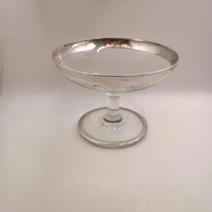 May include: A clear glass champagne coupe with a silver rim and base. The bowl of the glass has a textured surface. The stem is clear and the base is round.