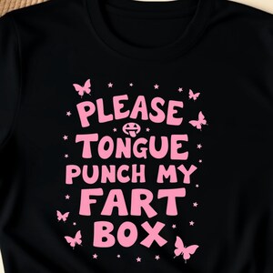 Könnte beinhalten: Schwarzes T-Shirt mit dem rosa Text "PLEASE TONGUE PUNCH MY FART BOX", verziert mit rosa Schmetterlingen und Sternen. Das T-Shirt ist unten geknotet. Das Wort "BLACK" befindet sich in der oberen rechten Ecke.