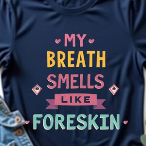 Puede incluir: Camiseta azul marino con el texto "MY BREATH SMELLS LIKE FORESKIN" en letras coloridas. Una chaqueta vaquera azul claro está doblada junto a la camiseta. Una ramita de hojas verdes está en el fondo.