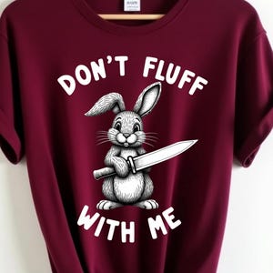 May include: Maroon t-shirt with the text "DON'T FLUFF WITH ME" and a graphic of a rabbit holding a knife. The shirt is hanging on a wooden hanger against a white background.