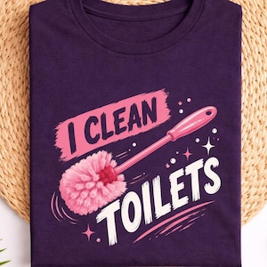 以下が含まれることがあります： ピンク色のトイレブラシのグラフィックと、白とピンクの文字で「I CLEAN TOILETS」と書かれたダークパープルのTシャツ。デザインには小さな星のアクセントとブラシのストロークが含まれており、折りたたまれた衣類の中央に配置されています。