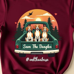 May include: Maroon t-shirt featuring a graphic of three beagles in a green truck bed with a sunset backdrop. The text "Save The Beagles" and "#endtheabuse" are displayed on the shirt. The shirt is knotted at the bottom.