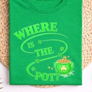 Könnte beinhalten: Irisch grünes T-Shirt mit dem Text "WHERE IS THE POT?" in hellgrüner Farbe. Das Design umfasst Kleeblätter und einen Goldtopf. Das Shirt wird auf einer gewebten Matte mit dekorativen Elementen präsentiert.
